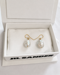 Paire de boucles d'oreilles Grainy Jil Sander en perle baroque blanche nacrée et crochet en laiton doré, présentées dans leur écrin signature blanc Jil Sander avec coussin en tissu blanc