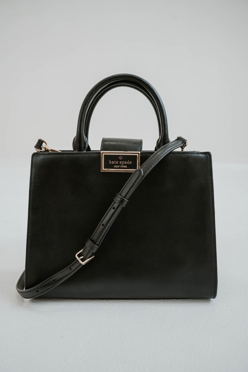Sac cartable Kate Spade Reegan en cuir lisse noir, silhouette structurée, fermeture magnétique à rabat, logo signature sur quincaillerie, bandoulière 22 pouces, compartiment zippé central, poche arrière intérieure _ vue de face