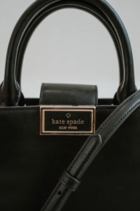 Sac cartable Kate Spade Reegan en cuir lisse noir, silhouette structurée, fermeture magnétique à rabat, logo signature sur quincaillerie, bandoulière 22 pouces, compartiment zippé central, poche arrière intérieure  - logo