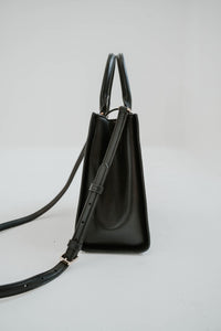 Sac cartable Kate Spade Reegan en cuir lisse noir, silhouette structurée, fermeture magnétique à rabat, logo signature sur quincaillerie, bandoulière 22 pouces, compartiment zippé central, poche arrière intérieure _ vue latérale