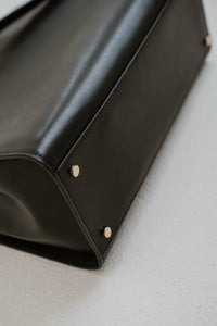 Sac cartable Kate Spade Reegan en cuir lisse noir, silhouette structurée, fermeture magnétique à rabat, logo signature sur quincaillerie, bandoulière 22 pouces, compartiment zippé central, poche arrière intérieure _ vue de dessous