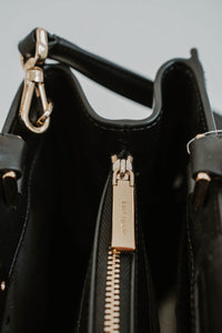 Sac cartable Kate Spade Reegan en cuir lisse noir, silhouette structurée, fermeture magnétique à rabat, logo signature sur quincaillerie, bandoulière 22 pouces, compartiment zippé central, poche arrière intérieure - glissière
